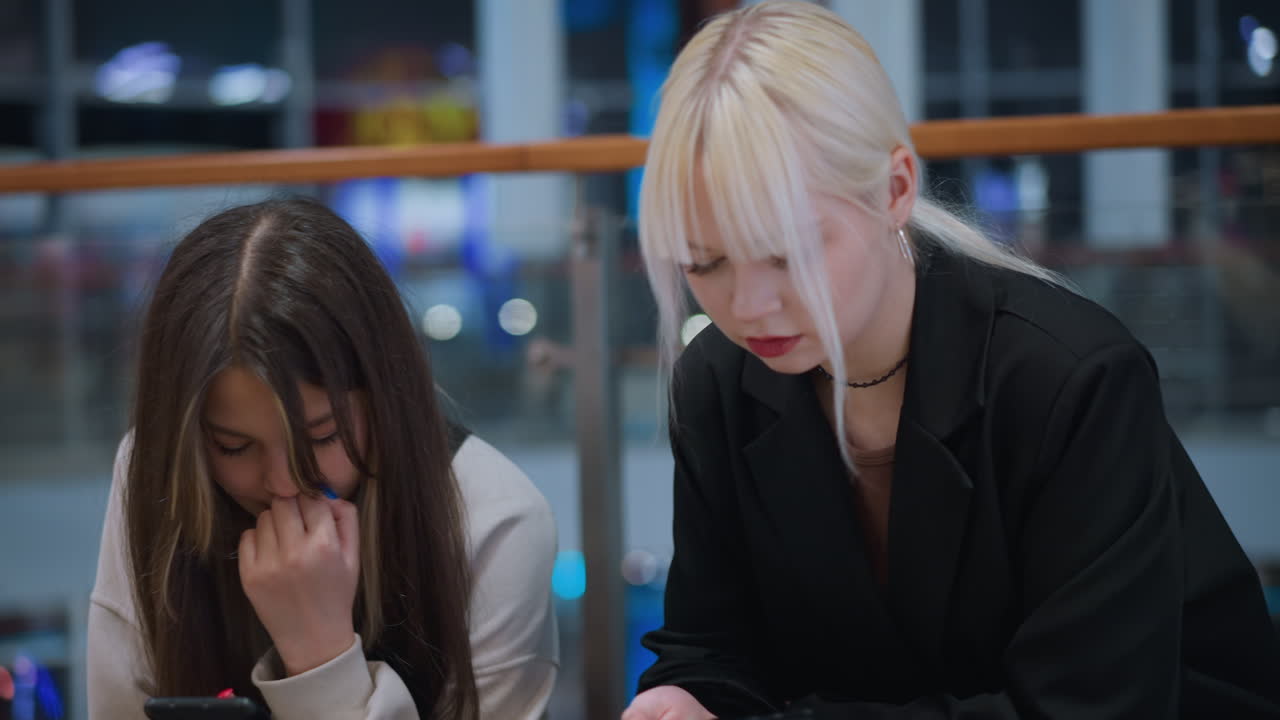 Close up of sisters sitting indoors holding phone with tired expression in modern mall showing casual lifestyle candid urban atmosphere portraying natural mood