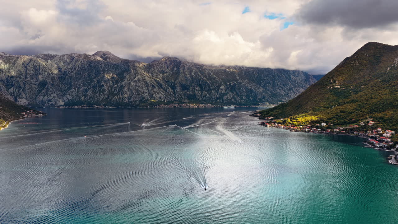 Sakin sularda hareket eden küçük teknelerin iz bıraktığı Kotor Körfezi'nin ve bulutlu gökyüzü altındaki heybetli kayalık dağların havadan drone ile çekilmiş görüntüsü