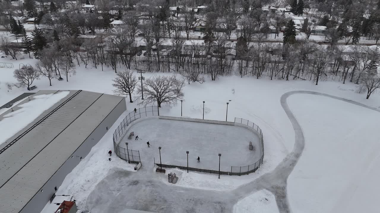 Letecký dron se přibližuje a obíhá doleva kolem venkovního kluziště ve Winnipegu, Manitoba, Kanada. Děti hrají hokej na zasněženém ledu s okolními domy a komunitním centrem.