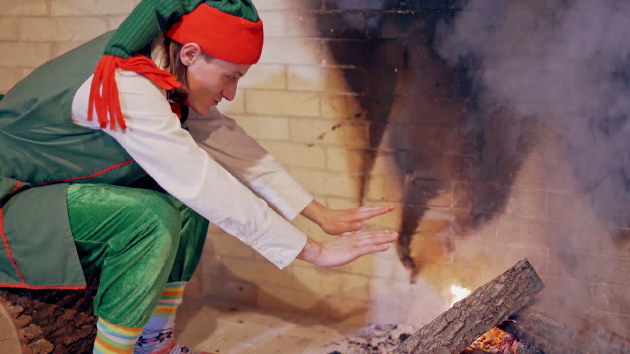 Elf is sitting near the fire and warming his cold hands in the house. Young man elf rubbing his cold hands near the fireplace. Fairy character elf is heating hands on Christmas.
