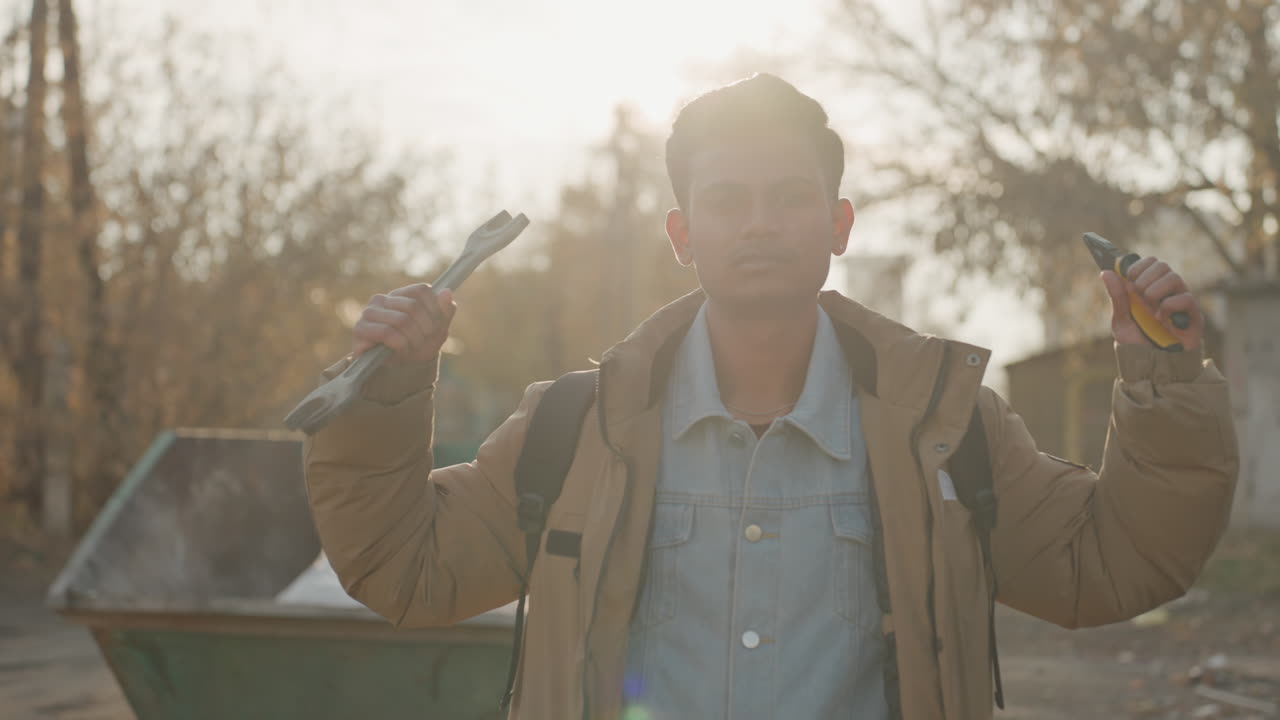 Uomo in controluce con attrezzi vicino a un cassonetto, in un vicolo autunnale, che rovista in cerca di materiale da recuperare, sollevando un cacciavite e delle pinze, con espressione contemplativa e un controluce caldo. Scena cinematografica suggestiva di riparazione, manutenzione urbana.