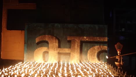 solo person lights row of hundreds of small candles on table using long red stick at night