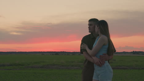 young couple standing closely in peaceful green field as woman affectionately rubs man side during golden sunset while both gaze softly into distance