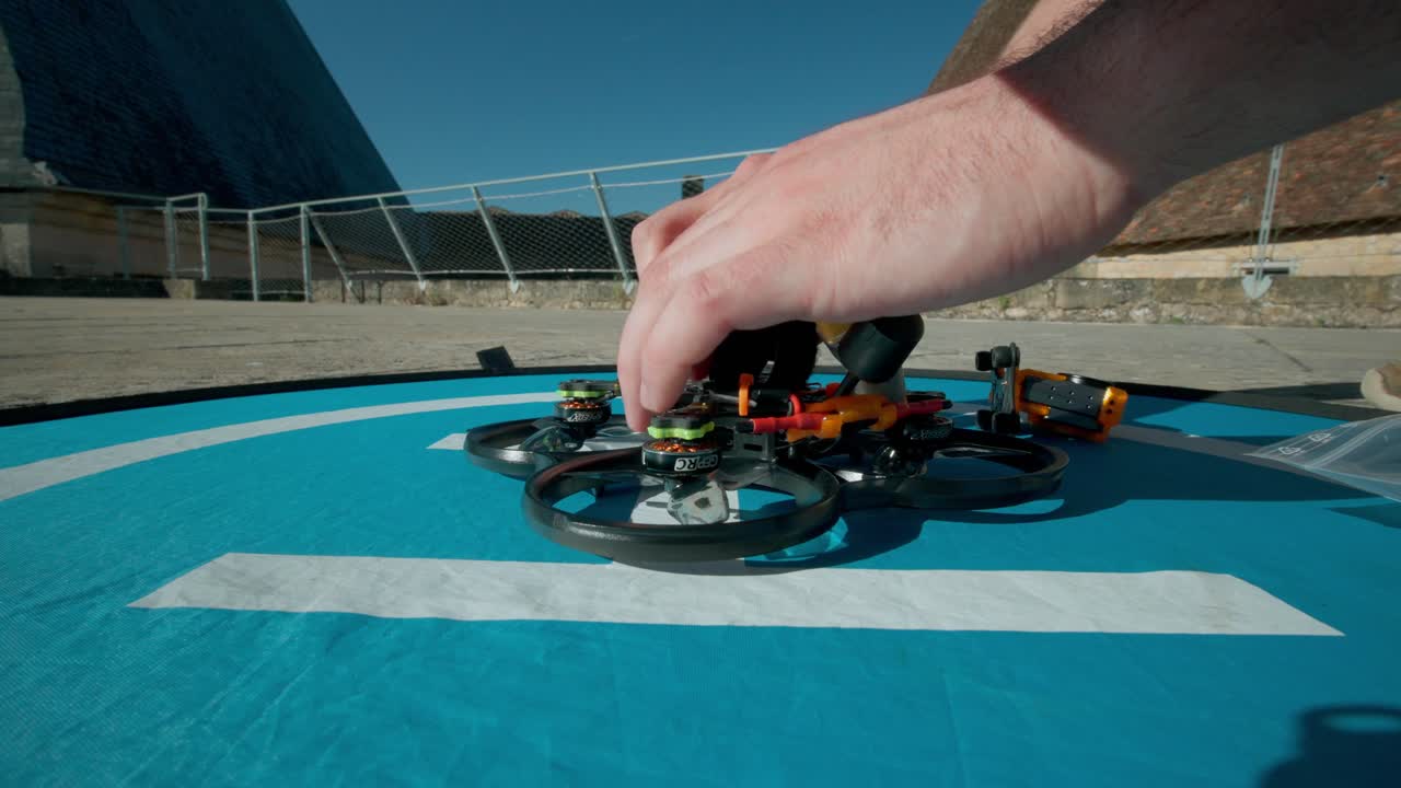 Mani che regolano il telaio e i componenti di un drone FPV nero posato su un tappetino di atterraggio blu prima del volo.