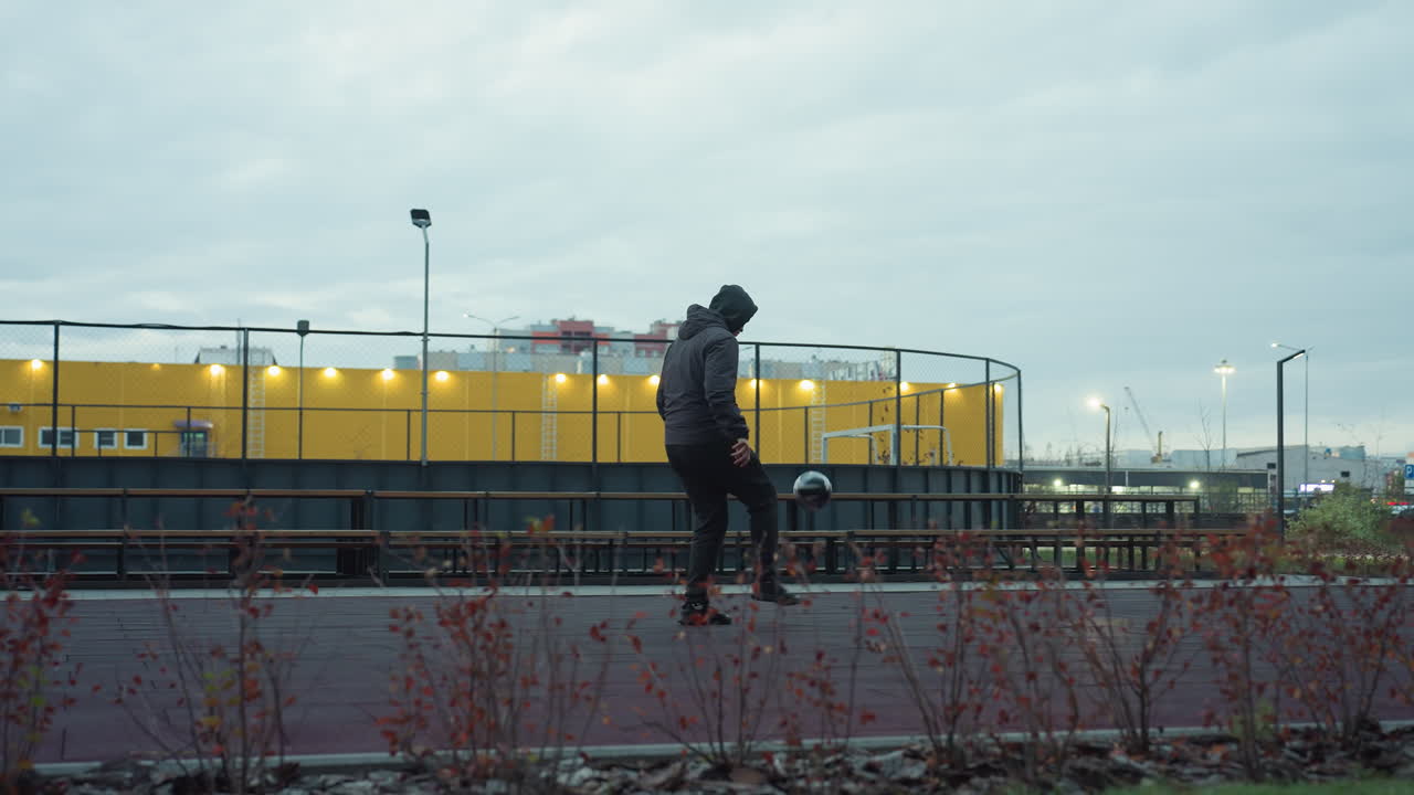 тренер умело жонглирует футболом на спортивной арене, окруженной городскими открытыми элементами, фон включает скамейки, игровую площадку, столбы освещения, зелень и городскую архитектуру