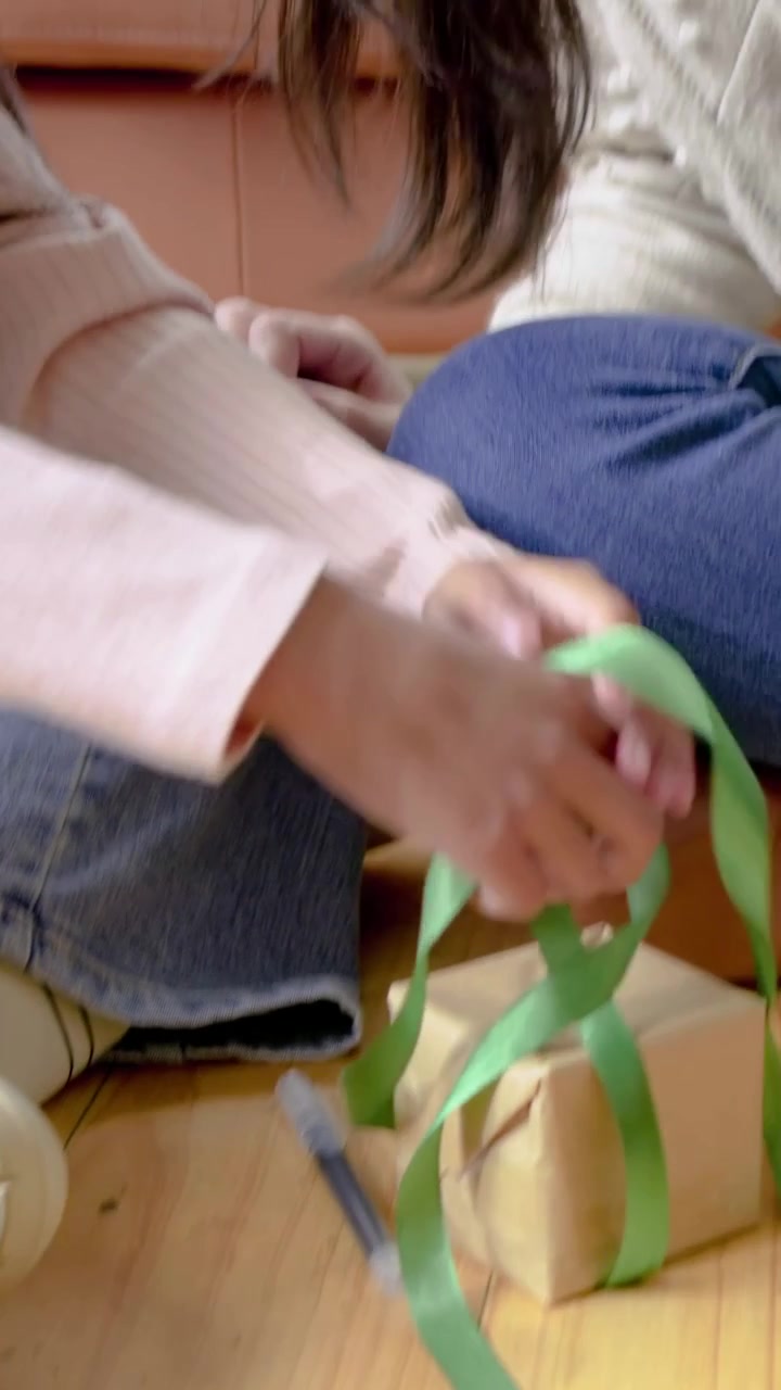 Video verticale: Bambino/a con top rosa che prende un nastro verde e impacchetta scatole di cartone kraft sul pavimento vicino al divano.