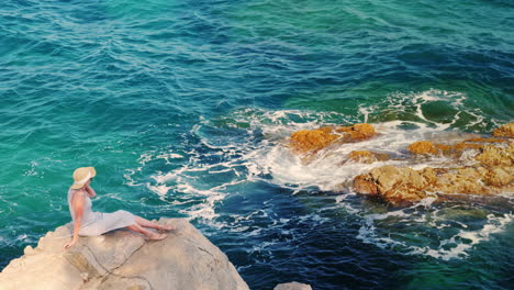 Woman-Tourist-In-Light-Dress-And-Hat-Sitting-On-A-Rock-Against-The-Sea