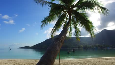 Palm-leaves-and-trunk-bent-over-white-sandy-beach-washed-by-calm-lagoon-reflecting-tropical-island-hill-with-lush-vegetation-in-Thailand