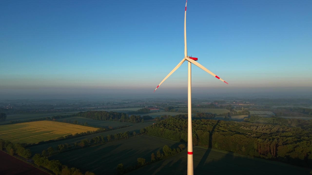 Singola turbina eolica che ruota sopra campi agricoli verdi e campi di colza gialli nell'Europa rurale durante il tramonto all'ora d'oro con cielo blu chiaro e orizzonte nebbioso sullo sfondo. Ripresa aerea in avvicinamento