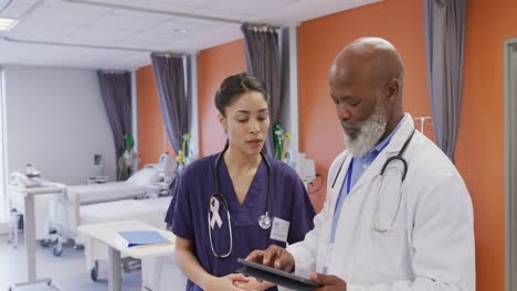 Two-diverse-male-and-female-doctors-with-tablet-wearing-cancer-awareness-ribbons-talking-at-hospital