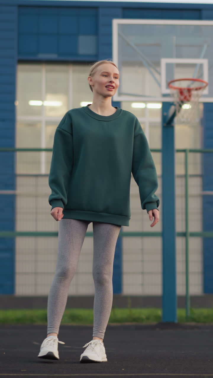 Giovane ragazza si allena nel servizio di pallavolo con un canestro da basket e un edificio sullo sfondo, dimostrando la tecnica sportiva su un campo all'aperto