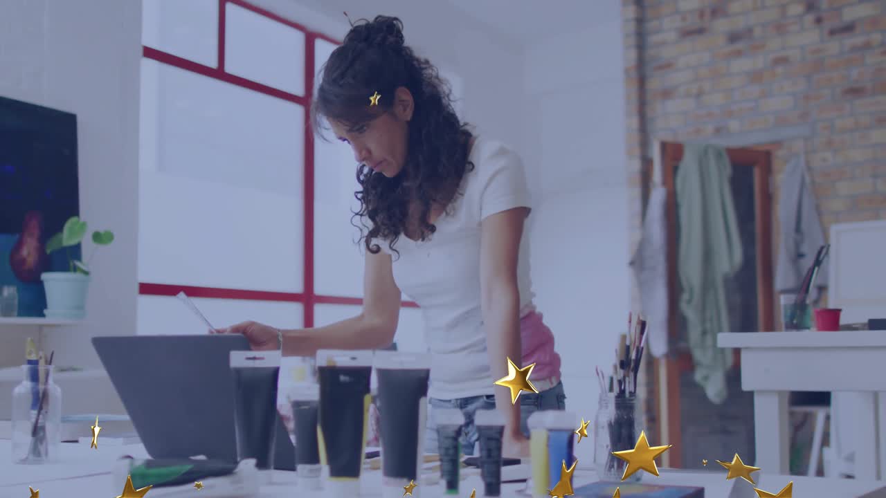 Woman leaning in studio studying laptop, lifting red brush, reviewing sketch while stars drifting