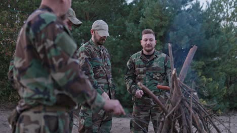 military men in camouflage clothing and cap stand outside around a campfire, talking, telling stories, frying sausages on sticks, resting after duty, hunting on base