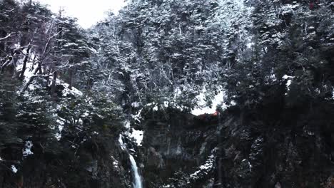 Toma-Aérea-Lenta-De-Un-Bosque-Montañoso-Nevado-Atravesado-Por-Una-Cascada