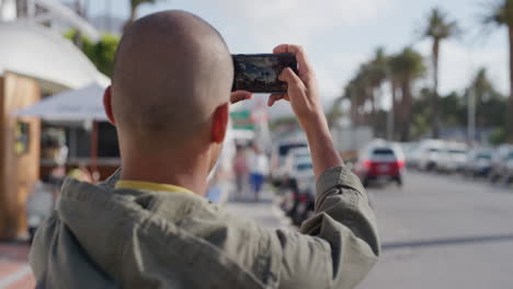 portrait of young bald man taking photo using smartphone on urban city beachfront enjoying sunny vibrant sightseeing vacation