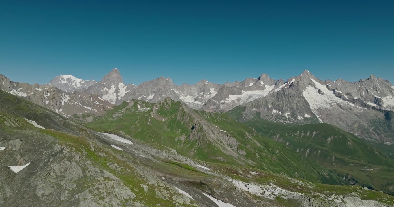 Drone terbang di atas medan dramatis Colle del Gran San Bernardo, memperlihatkan lanskap pegunungan Alpen yang terjal, puncak-puncak bersalju, dan langit musim panas biru jernih di perbatasan Swiss-Italia