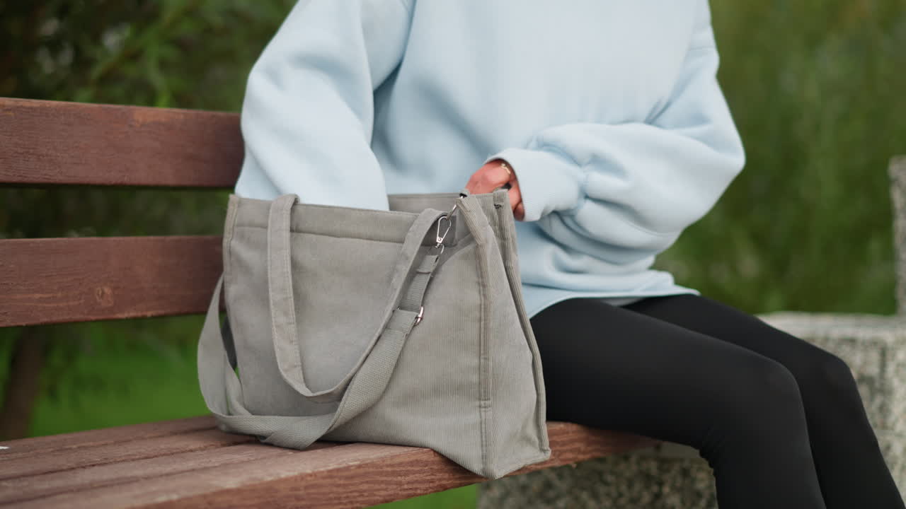 Partial view of lady sitting on concrete bench in casual wear, pulling water bottle out from her ash-colored handbag, calm outdoor setting, ideal for hydration, relaxation, and wellness moments