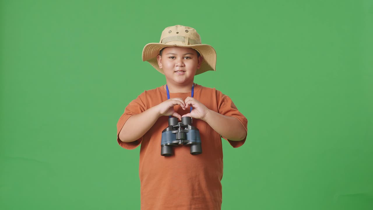 niño turista asiático con un sombrero y binoculares haciendo un gesto en forma de corazón mientras está de pie en el fondo de la pantalla verde. niño investigador examina algo, concepto de aventura de turismo de viaje
