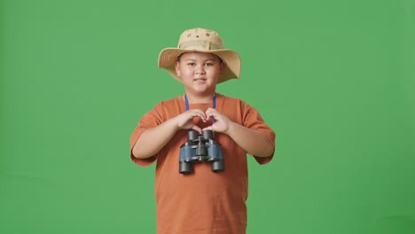 asian tourist boy with a hat and binoculars making heart shape gesture while standing on the green screen background. boy researcher examines something, travel tourism adventure concept