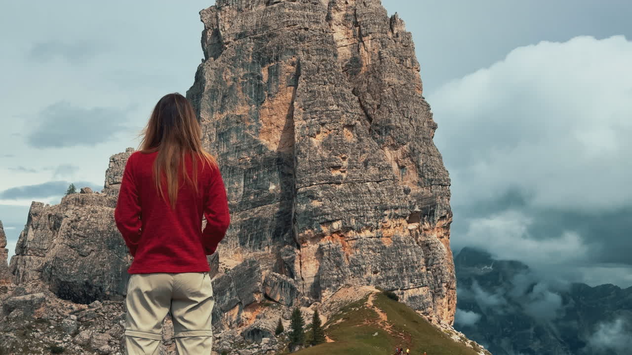 Mountain hiker standing on rocky terrain, gazing at dramatic Cinque Torri peaks stretching across scenic Dolomites landscape with cloudy alpine sky