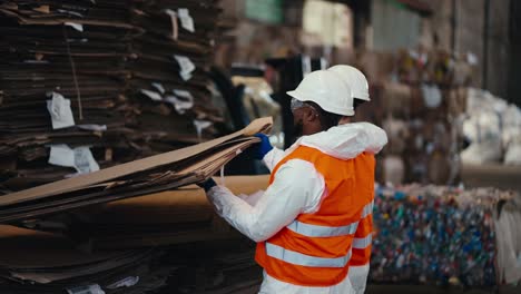 un hombre de piel negra en un uniforme de protección blanco en un chaleco naranja junto con su colega un hombre con barba levanta un gran estante de cartón en una planta de reciclaje y clasificación de residuos para papel y plástico