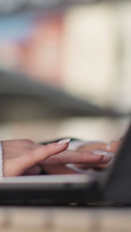 primer plano de las manos de la dama con uñas pulidas y anillos delicados escribiendo en el teclado de la computadora portátil al aire libre, colocado en una mesa de madera con fondo borroso