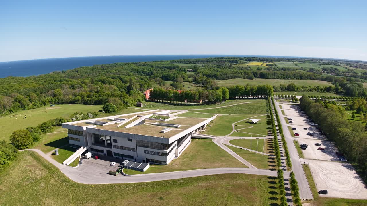 Aerial view of a sleek, modern building situated in a green landscape with open fields and a symmetrical road layout. Captured on a clear sunny day