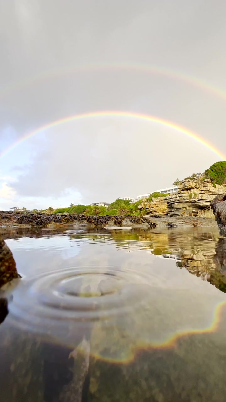 Verticale. Doppio arcobaleno riflesso in una pozza di roccia oceanica mentre leggere gocce di pioggia increspano l'acqua.