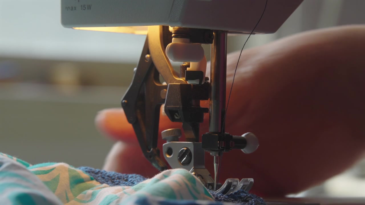 Close up of sewing machine needle stitching as woman feeds frilly blue fabric through, indoor setting