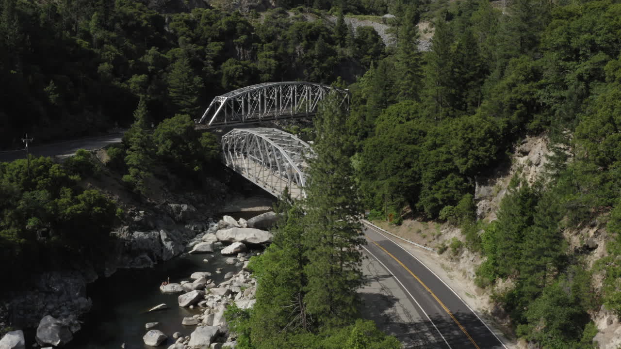 Cinematic shot of a silver car driving on rural road between mountains and pine trees, passing over metal arched bridge, tracking shot, aerial