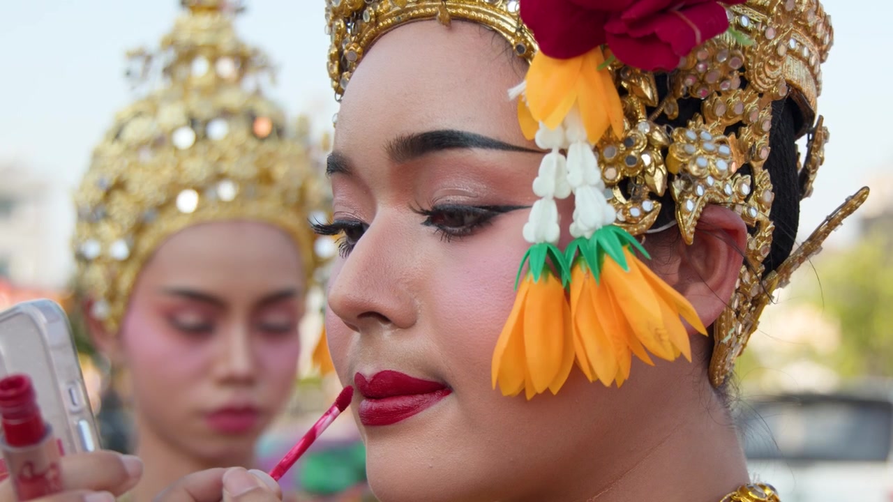 Cận cảnh góc nghiêng một vũ công Thái Lan trong trang phục truyền thống đang thoa son môi đỏ ngoài trời.