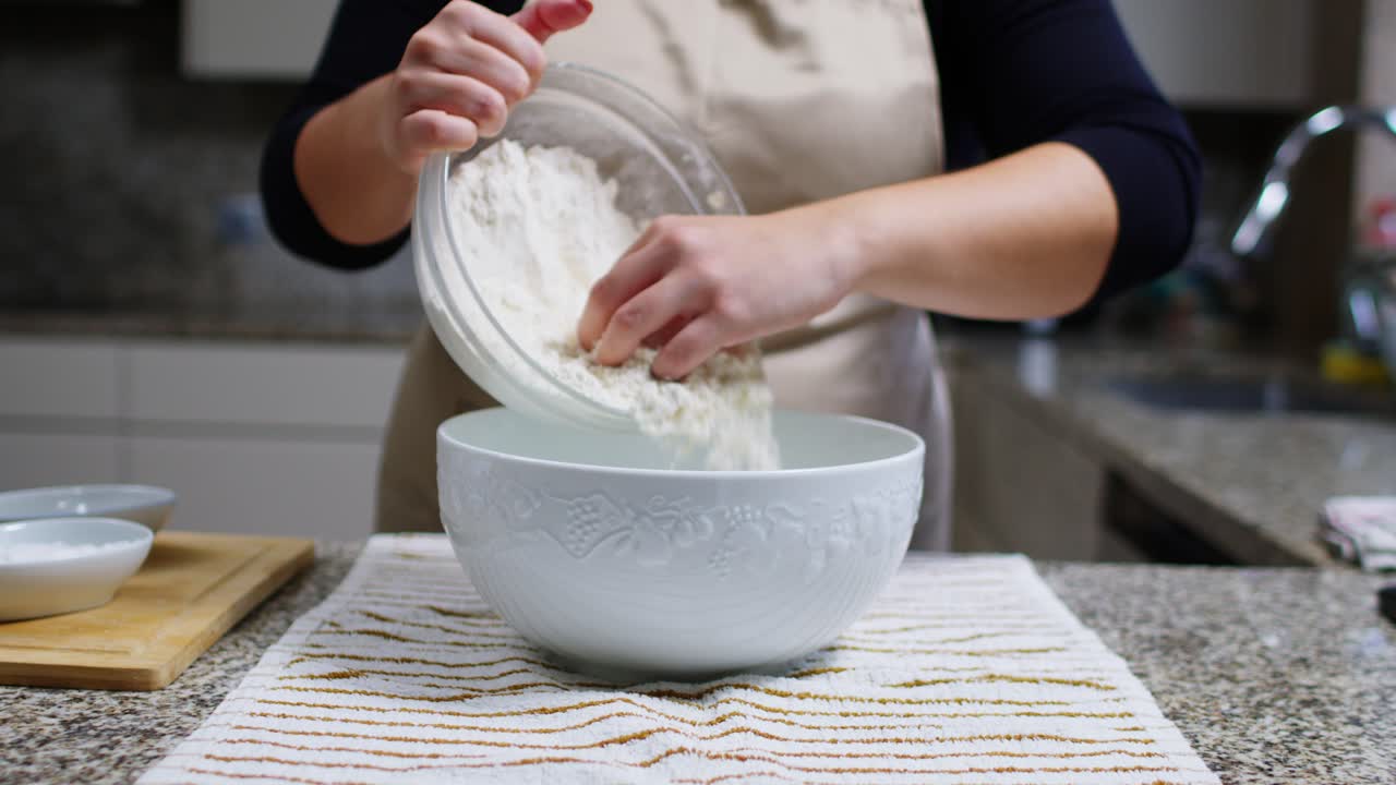 Woman pours white flour into a ceramic bowl from another bowl in a kitchen.