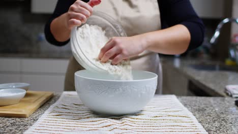 una mujer vierte harina blanca en un cuenco de cerámica de otro cuenco en una cocina.