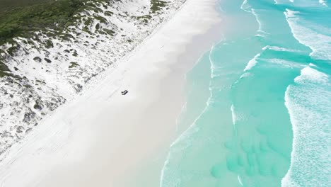 Excellent-Aerial-Shot-Of-A-Van-Driving-On-The-White-Sands-Of-Wharton-Bay-As-Clear-Blue-Water-Laps-The-Shore-In-Esperance,-Australia