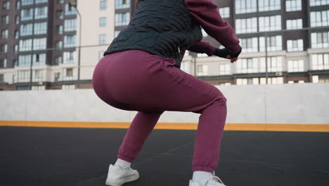 back view woman squatting on round sport court at sunset with warm light on tall buildings creating urban workout silhouette emphasizing strength stability balance in dusk city setting