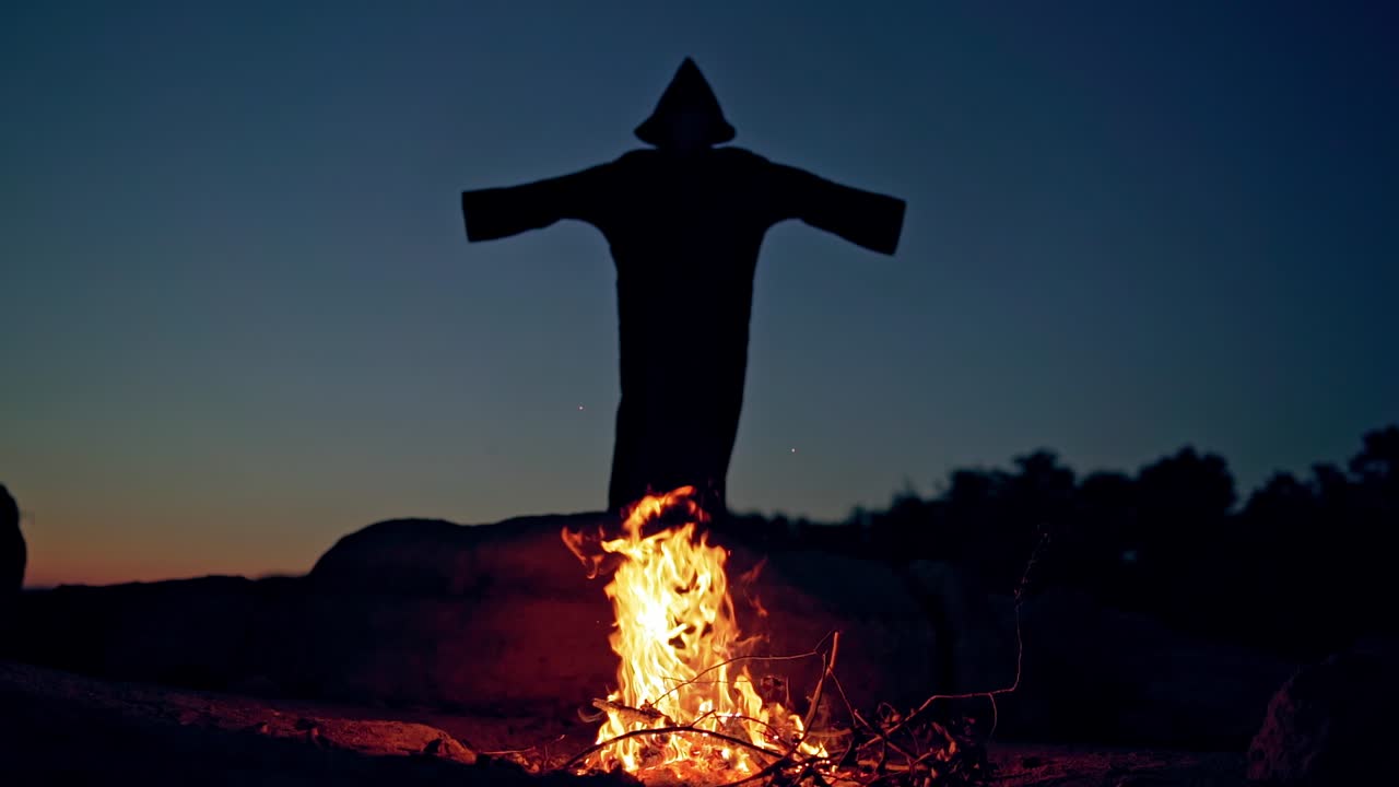 Black death behind the fire flame outdoors. Scary witch in black costume waving arms making fear on the background of burning fire. Halloween costume.