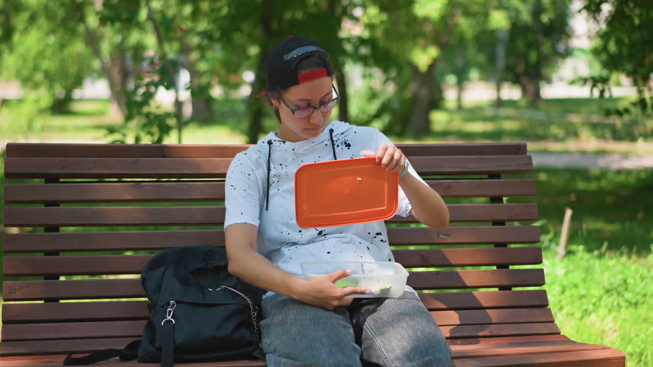 Youth enjoys outdoor meal, Casual teen eats lunch outdoors, Young individual has relaxing midday meal outside, Juvenile person seated on park bench consumes lunch peacefully outdoors