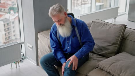 An-elderly-man-with-gray-hair-and-a-bushy-beard-massages-his-knee-while-sitting-on-a-brown-sofa-in-a-modern-apartment.-An-elderly-man-stretches-his-knee-after-a-long-walk-in-a-modern-apartment-with-large-windows