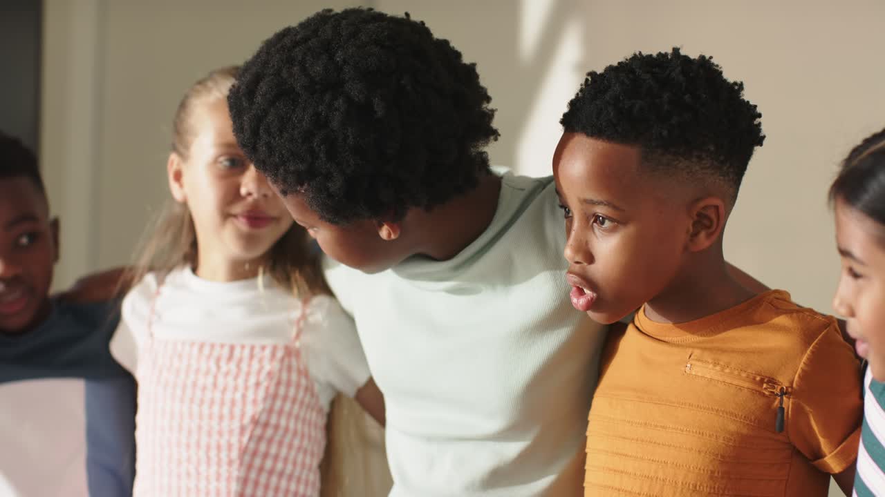 Crianças diversas de braços dados, conversando após uma deixa em sala de aula, menina de vestido rosa fazendo uma apresentação