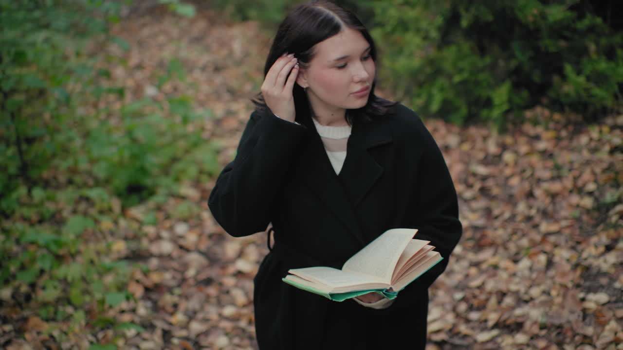 Vista desde arriba de una chica guapa con abrigo de otoño hojeando un libro mientras pasea por un sendero forestal, mirada tranquila alrededor, follaje bokeh suave, momento de lectura agradable, hojas que crujen bajo sus pies