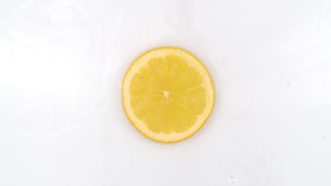 On a white background a splash of water falls on a slice of lemon in slow motion.