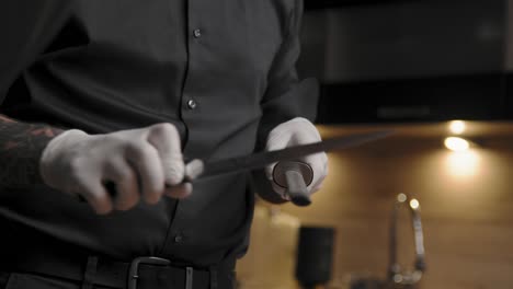 Young-professional-male-chef-in-an-elegant-black-shirt-with-tattoos-taking-a-knife-from-a-block-and-sharpens-it