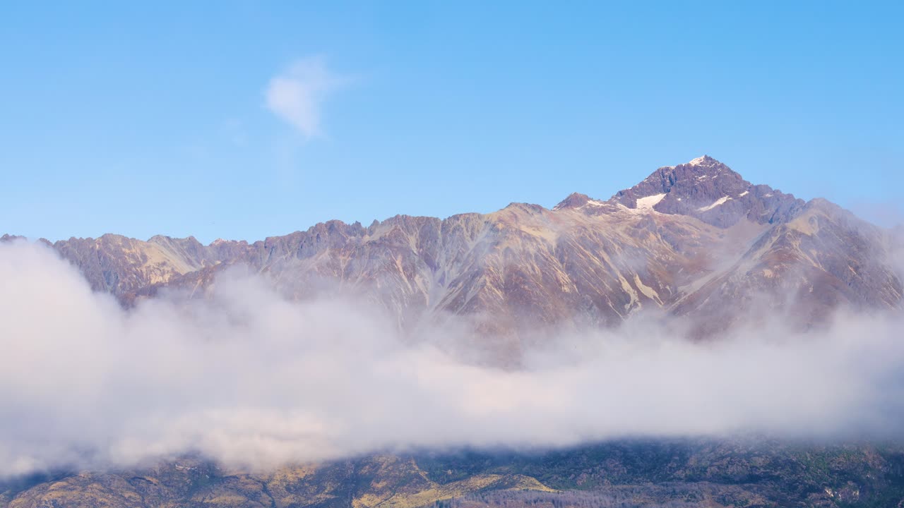 Mountain peaks rise above drifting fog under a clear blue sky in Queenstown, New Zealand. Captured in serene natural lighting