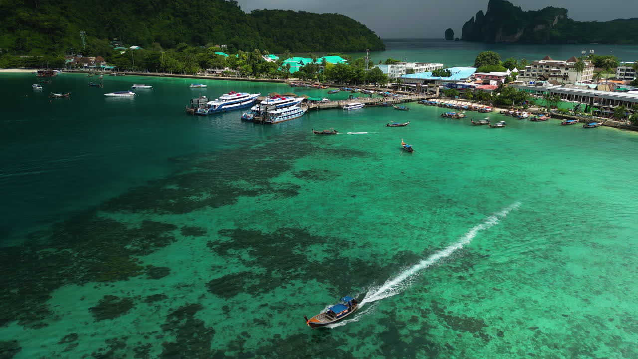 Stunning Aerial Tracking Shot Of A Long Tail Boat Water Taxi Departing The Busy Seaport Village Of Koh Phi Phi, One Of Thailand's Most Visited Islands