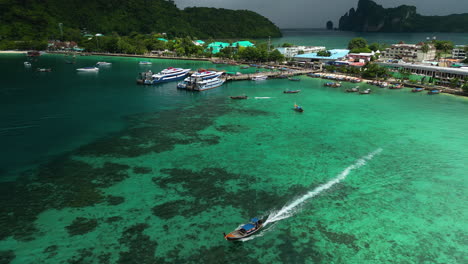 Stunning-Aerial-Tracking-Shot-Of-A-Long-Tail-Boat-Water-Taxi-Departing-The-Busy-Seaport-Village-Of-Koh-Phi-Phi,-One-Of-Thailand's-Most-Visited-Islands