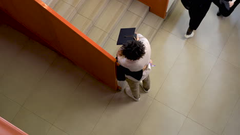 top view of a group of preschool students in cap and gown running down the stairs holding their graduation diplomas while one of them hugging the teacher