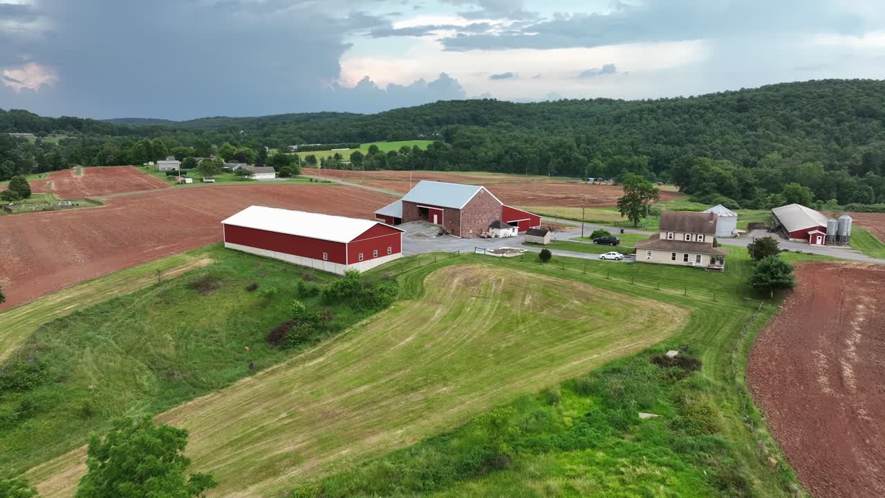 Aerial Approaching shot of American farm in rural area with red brick barn building in summer. Large property with stable and Thatched roof farm house