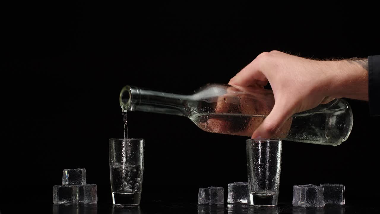barman vertiendo vodka de la botella en dos copas con cubos de hielo en fondo negro