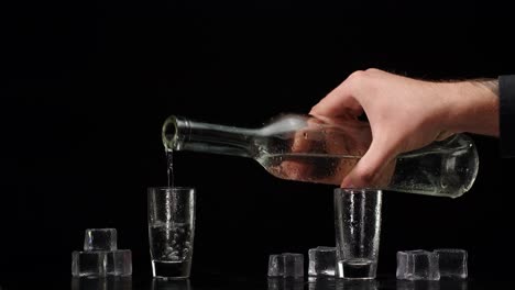 Bartender-pouring-up-vodka-from-bottle-into-two-shots-glasses-with-ice-cubes-on-black-background