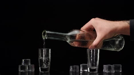bartender pouring up vodka from bottle into two shots glasses with ice cubes on black background